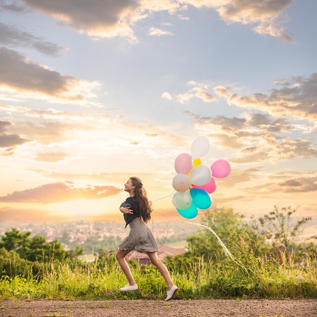 Menina bonita com grandes balões coloridos andando nas colinas perto da cidade no pôr do sol