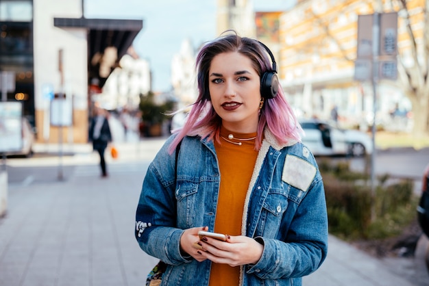 Menina bonita com música de escuta do cabelo tingido em fones de ouvido na rua da cidade.