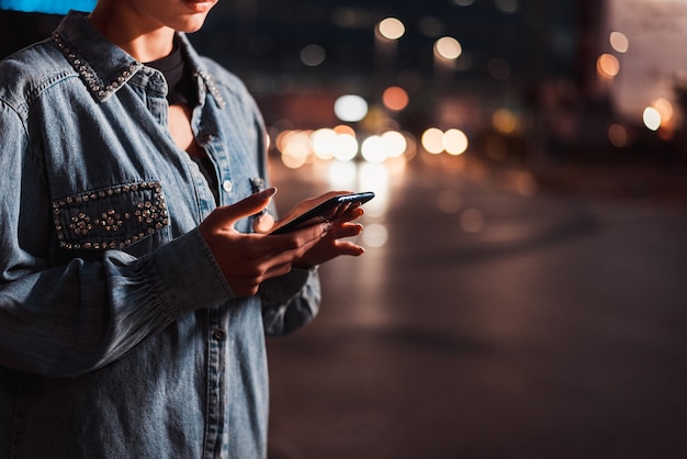 Menina bonita com roupas elegantes segurando um smartphone ao ar livre à noite, cidade iluminada