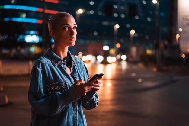 Menina bonita com roupas elegantes segurando um smartphone ao ar livre à noite, cidade iluminada