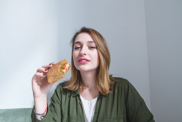 Menina bonita com sanduíche nas mãos dela olha sorrisos de camerand. mulher tem café da manhã fast-food