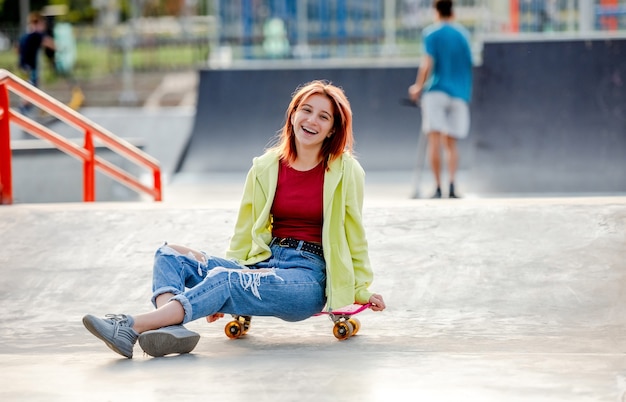 Menina bonita com skate sentado e sorrindo. Patinadora adolescente perto da rampa do parque