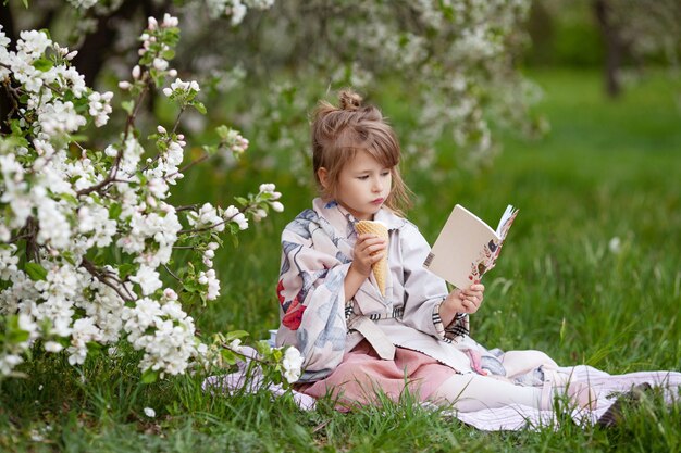 Menina bonita com sorvete, lendo um livro no parque. criança ao ar livre em um jardim florido de primavera