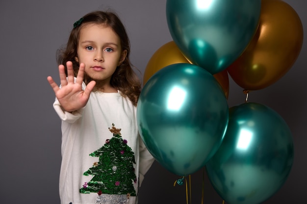 Menina bonita com suéter de Natal fica perto de belas bolas de ar metálicas e douradas verdes brilhantes e mostra um sinal de STOP, gesticulando com o dedo, isolado sobre um fundo cinza com espaço de cópia para anúncio