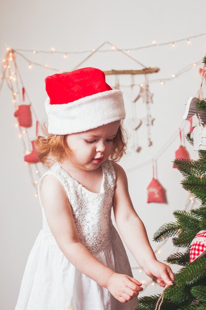 Menina bonita com um boné de Papai Noel decorando a árvore de Natal