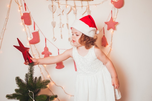 Menina bonita com um boné do Papai Noel pendurando uma estrela no topo da árvore de Natal