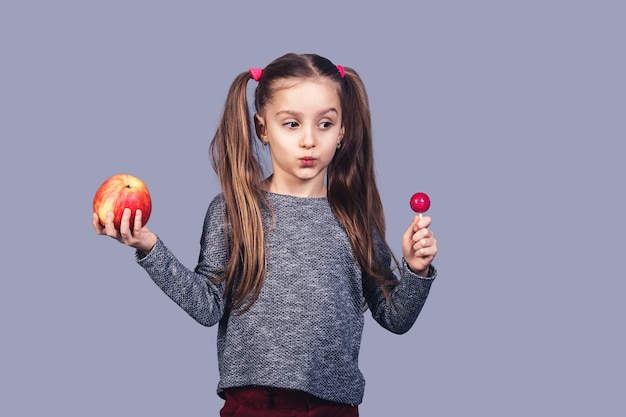 Menina bonita com um doce e uma maçã nas mãos. Escolha do conceito entre comida saudável e junk food. isolado na superfície cinza