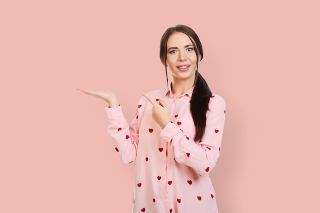 Menina bonita com um sorriso em um fundo rosa em uma camisa rosa com corações vermelhos aponta os dedos para o local para ofertas de promoções ou publicidade