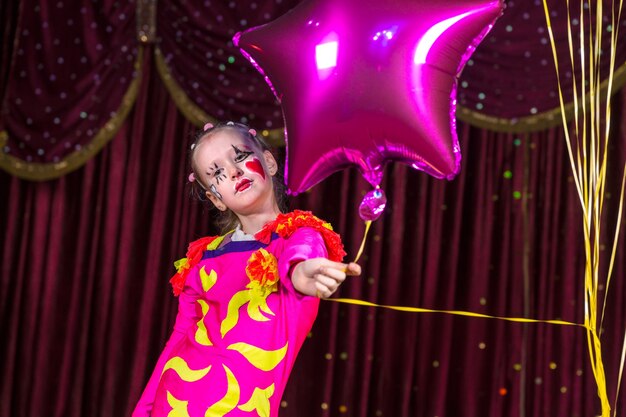 Menina bonita com um traje festivo em rosa e amarelo de cores vivas segurando um balão de alumínio em forma de estrela para a câmera enquanto sobe no palco