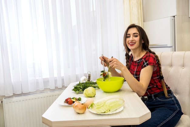 Menina bonita comendo salada de legumes frescos enquanto sorrindo e sentado na cozinha em casa.