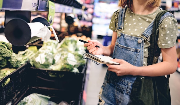 Menina bonita comprando com lista de compras no supermercado e procurando produtos nas prateleiras bonitas