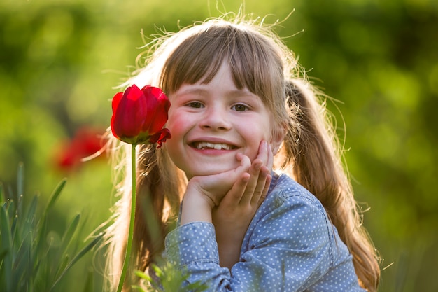 Menina bonita criança sorridente com olhos cinzentos e cabelos longos com flor tulipa vermelha brilhante no fundo desfocado ensolarado bokeh verde. Amor ao conceito de natureza.