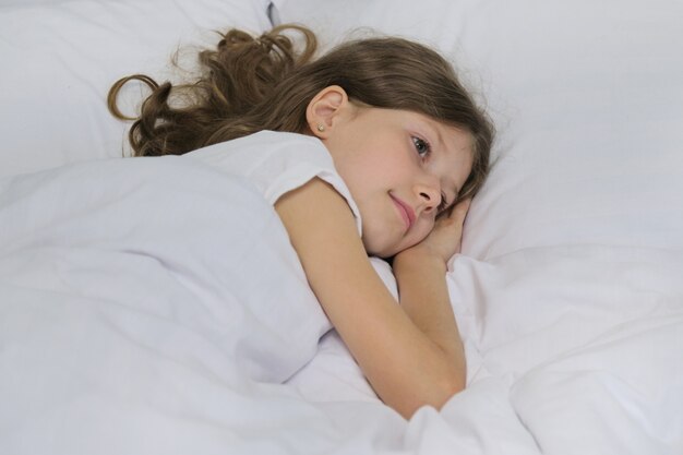 Menina bonita criança sorridente, deitado sobre um travesseiro, cama branca, rosto de close-up