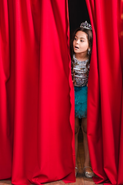 Menina bonita curiosa a espreitar da cortina vermelha no palco