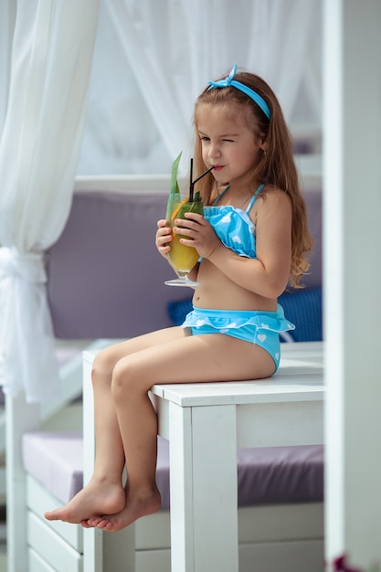 Menina bonita de 5 anos com cabelos escuros em um maiô azul de bolinhas e óculos de sol bebe um coquetel nas férias de verão à beira da piscina.