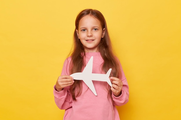 Menina bonita de cabelos castanhos positiva vestindo camisola rosa segurando avião de papel branco isolado sobre fundo amarelo olhando para a câmera de publicidade da companhia aérea