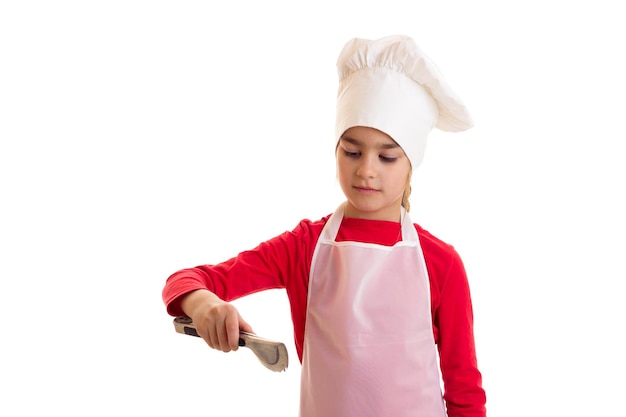 Menina bonita de camisa vermelha com avental branco e chapéu segurando uma pinça de cozinha em estúdio