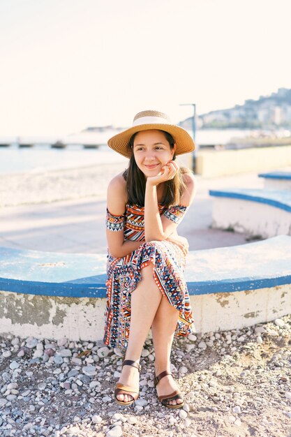 Menina bonita de chapéu e vestido sentada em um banco em uma praia de seixos