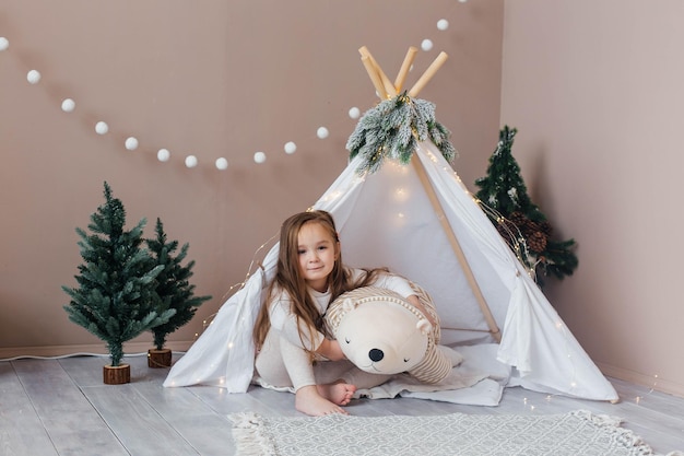 Menina bonita de pijama branco brinca com um ursinho de pelúcia perto do quarto das crianças da tenda com c