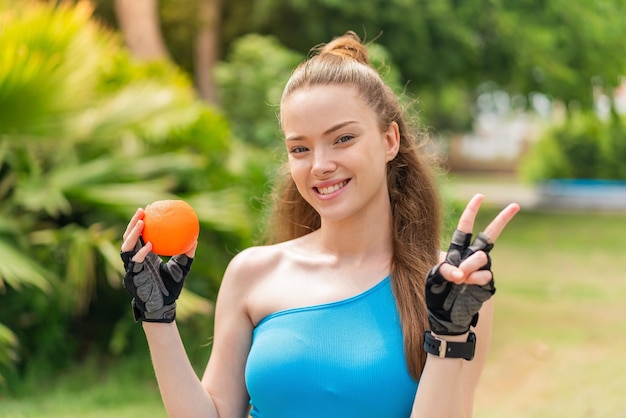 Menina bonita do esporte segurando uma laranja ao ar livre sorrindo e mostrando sinal de vitória