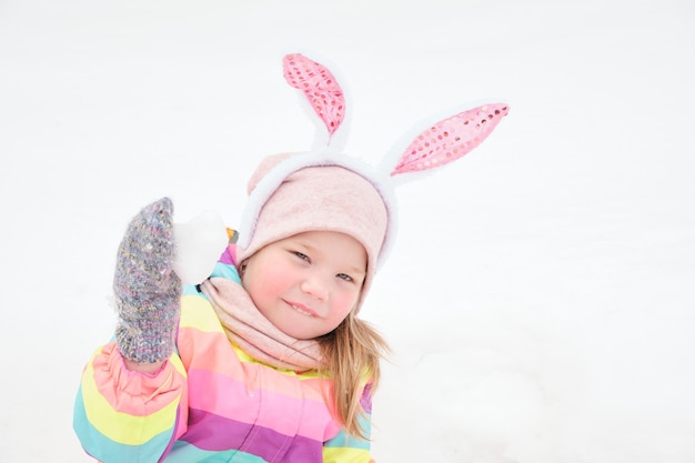 Menina bonita e alegre em idade pré-escolar com orelhas de coelho caranal na cabeça brinca com um coração feito de neve em abril