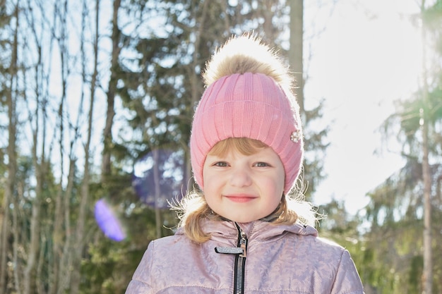 Menina bonita e alegre em uma floresta de neve brilhante sol de primavera neve na primavera uma garota com um chapéu rosa aperta os olhos