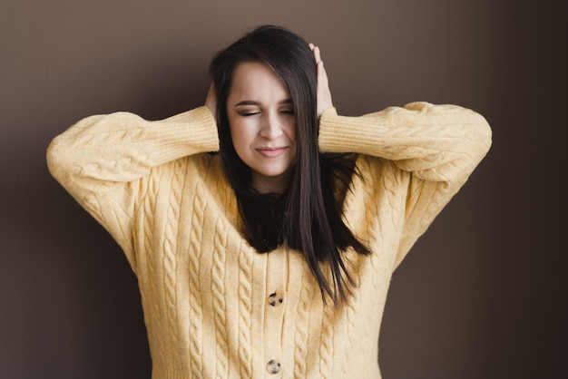 Menina bonita e bonita em um suéter amarelo em um fundo marrom isolado, cobrindo os ouvidos