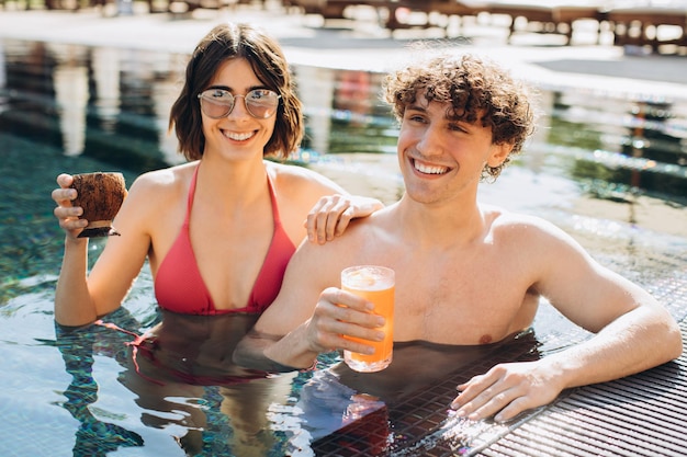 Menina bonita e cara encaracolado bebendo coquetéis e se divertindo à beira da piscina no verão