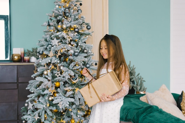 Menina bonita e feliz abre um presente de Natal perto de uma árvore de Natal festiva