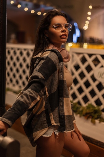 Menina bonita elegante com óculos da moda e fones de ouvido em roupas casuais de verão da moda com shorts de camisa xadrez e um top caminha na cidade à noite