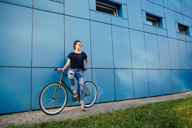 Menina bonita elegante em pé com bicicleta vintage e desviar o olhar, perto da parede azul, aproveitando o tempo de lazer ao ar livre.