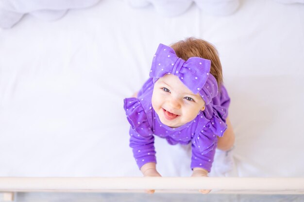 Menina bonita em um berço está segurando ao lado em roupas lilás e com um laço na cabeça bebê engraçado vista superior conceito de produtos infantis