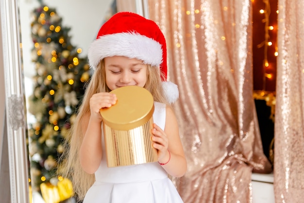 Menina bonita em um lindo vestido branco, com um chapéu de Papai Noel abre um presente de Natal.