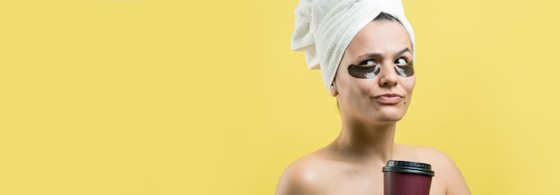 Menina bonita em uma toalha branca na cabeça usa adesivos de gel de colágeno sob os olhos Máscara sob o rosto de tratamento dos olhos A garota tem um copo de café nas mãos