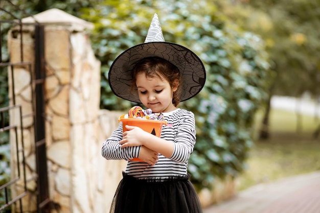 Menina bonita fantasiada de bruxa segurando um balde de abóbora jackolantern com doces e doces Kid truque ou tratamento no feriado de Halloween