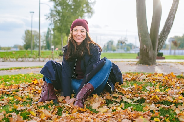 Menina bonita feliz senta-se no outono em folhas amarelas no gramado da cidade.