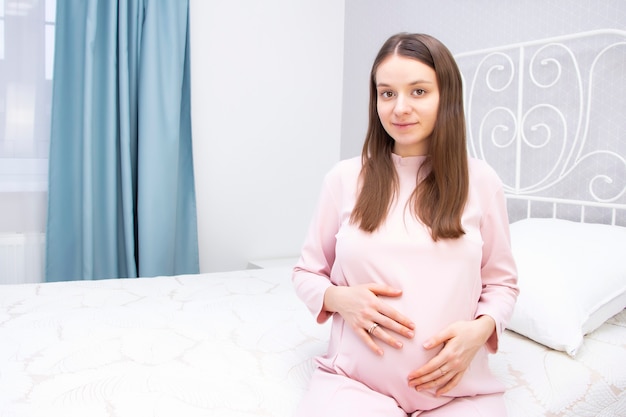 Menina bonita grávida está sentada na cama e as mãos estão na barriga. conceito de gravidez.