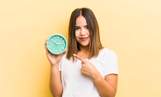 Menina bonita hispânica sorrindo alegremente se sentindo feliz e apontando para o conceito de despertador lateral