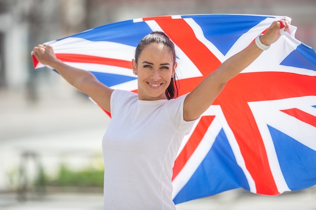 Menina bonita ina t-shirt branca segura uma bandeira da Grã-Bretanha atrás dela, sorrindo para a câmera.