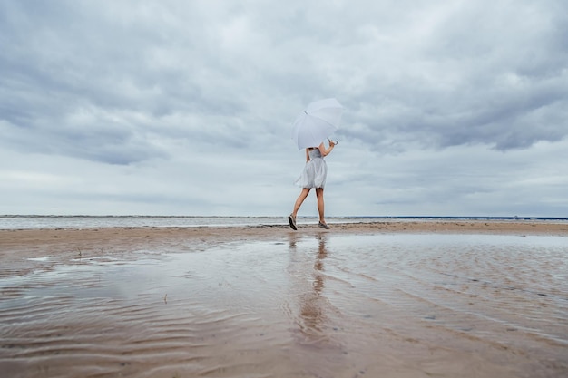 Menina bonita irreconhecível de vestido cinza com guarda-chuva branco corre ao longo do mar