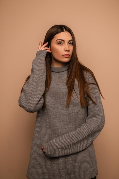 Menina bonita jovem com beleza natural em um suéter de malha elegante fica perto de fundo bege no estúdio. retrato de mulher