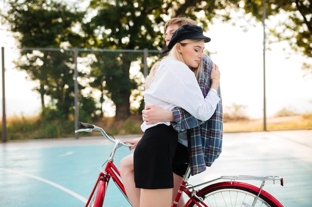 Menina bonita jovem com cabelo loiro sonhadoramente fechando os olhos enquanto abraçava o menino na bicicleta vermelha na quadra de basquete no parque Retrato de lindo casal abraçando um ao outro