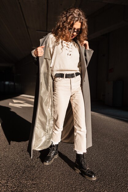 Menina bonita jovem com cabelos cacheados em roupas da moda com um casaco vintage, óculos escuros, suéter, calças e botas caminha pela cidade em um dia ensolarado