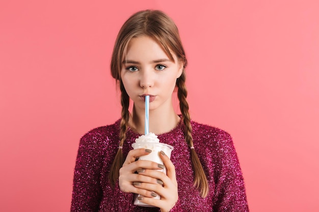 Menina bonita jovem com duas tranças no suéter bebendo milk-shake olhando pensativamente na câmera enquanto passa o tempo sobre fundo rosa