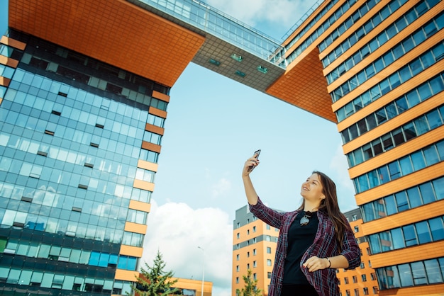 Menina bonita jovem em uma camisa leva selfie em seu smartphone e sorrisos. morena de cabelos compridos com um telefone em uma rua da cidade, close-up.