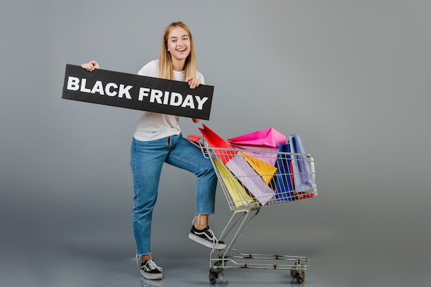 Menina bonita jovem sorridente com sinal preto de sexta-feira e carrinho de mão cheio de sacolas coloridas