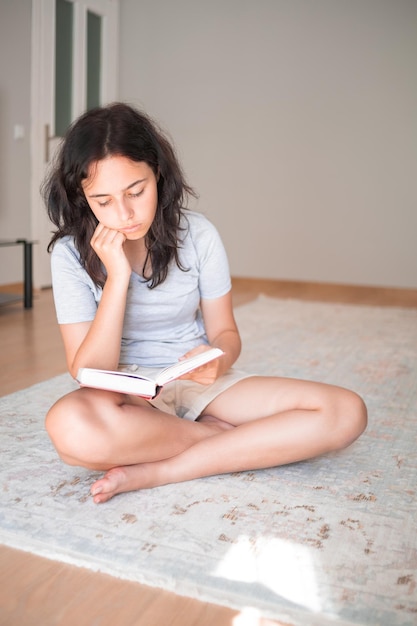 Menina bonita lendo livro de papel enquanto está sentado no tapete em casa