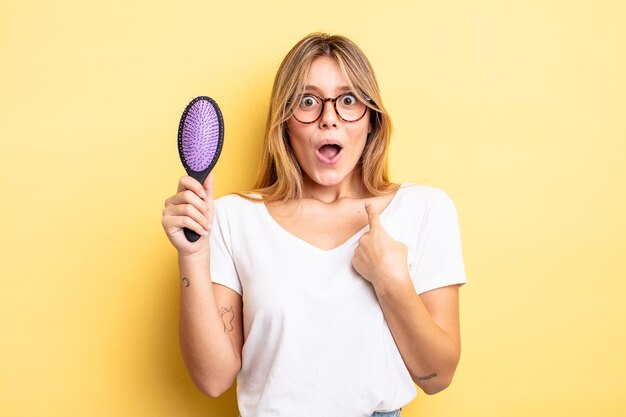 Menina bonita loira parecendo chocada e surpresa com a boca aberta apontando para o conceito de escova de cabelo próprio