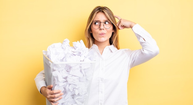 Menina bonita loira se sentindo perplexa e confusa, coçando a cabeça. conceito de lixo de bolas de papel