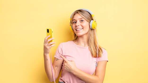 Menina bonita loira sorrindo alegremente, sentindo-se feliz e apontando para o lado. ouvir conceito de música